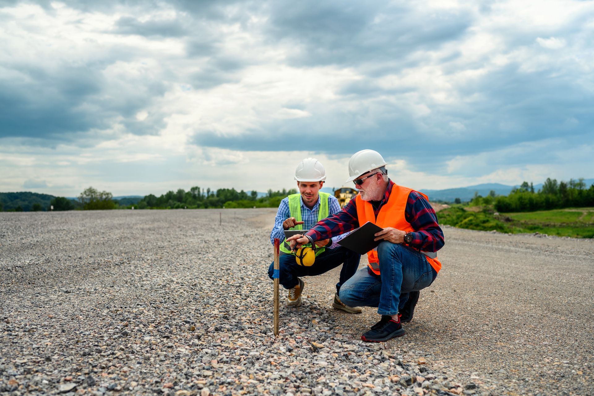 Two construction workers in hard hats and vests examine a gravel surface outdoors under a cloudy sky.