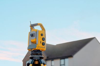 Yellow total station surveying equipment on a tripod, set up in front of a house with a blue sky background.