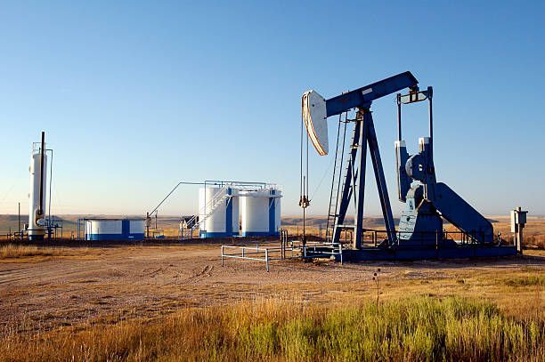 An oil pumpjack in a dry, grassy field with storage tanks, under a blue sky.