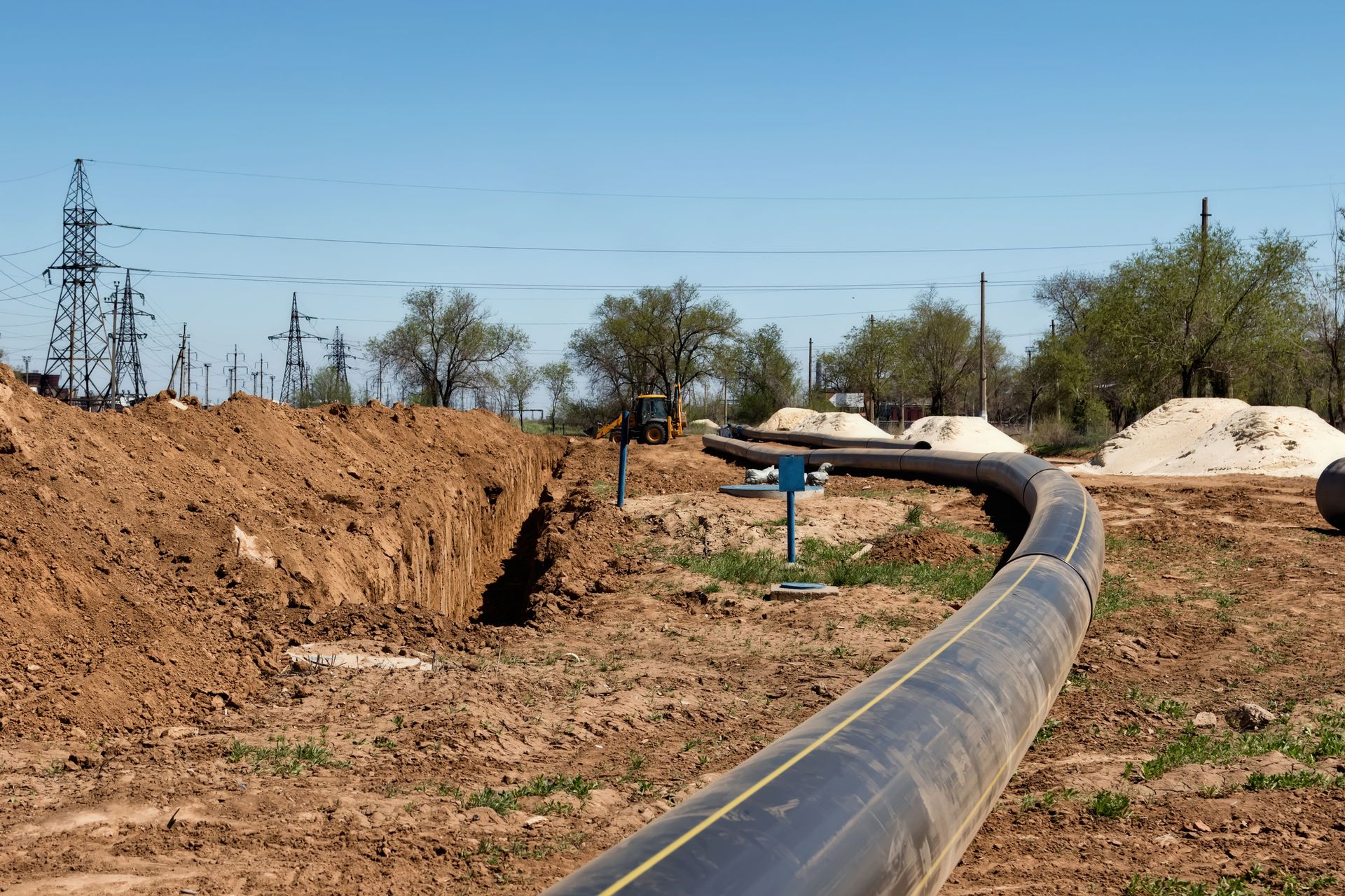 A long, black pipeline curving across a construction site with a trench, dirt piles, and blue markers under a blue sky.