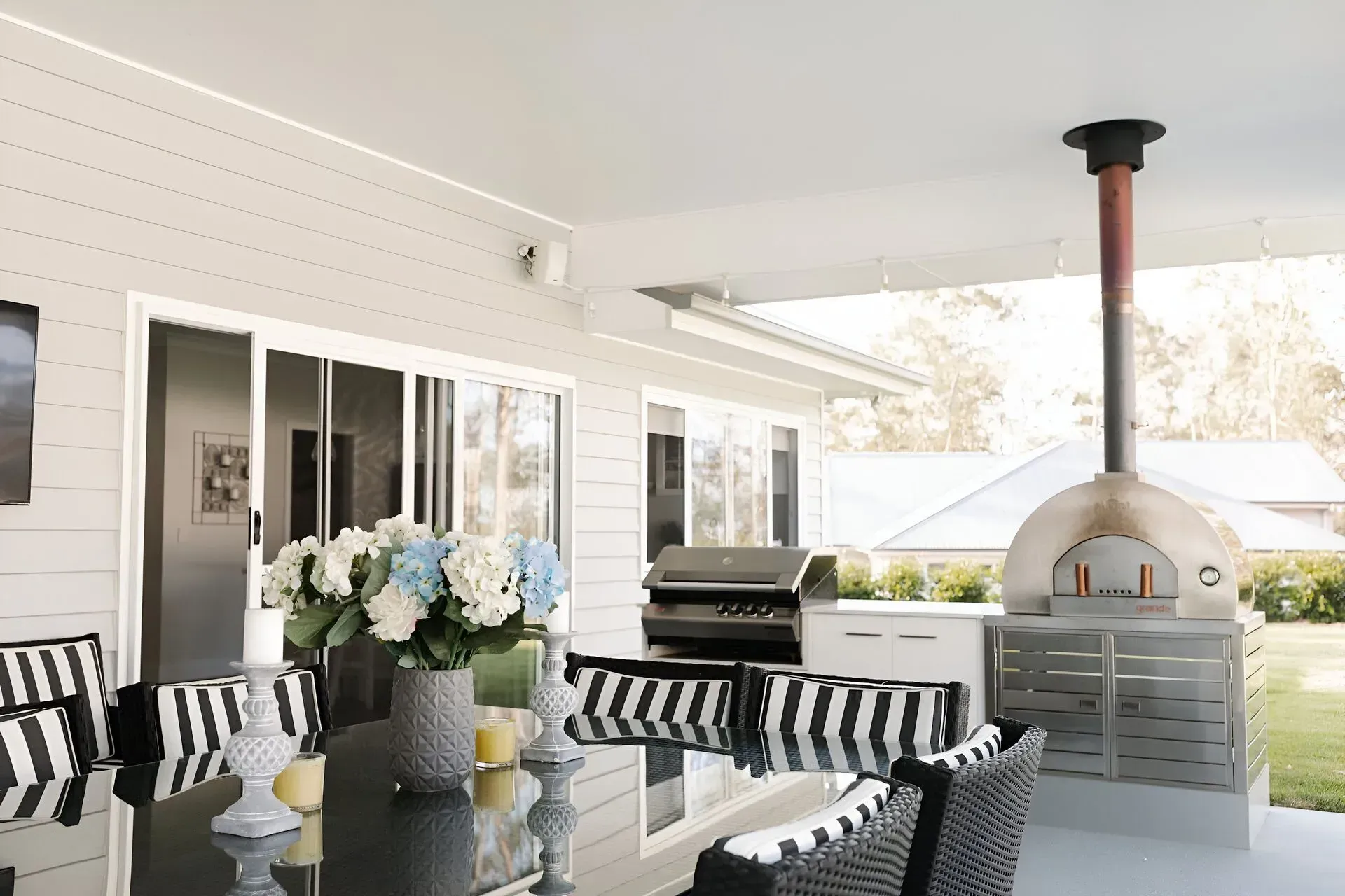 Outdoor patio with a table, striped chairs, a grill, and a pizza oven under a covered roof. — Michael Duncan Constructions Pty Ltd in Robina, QLD