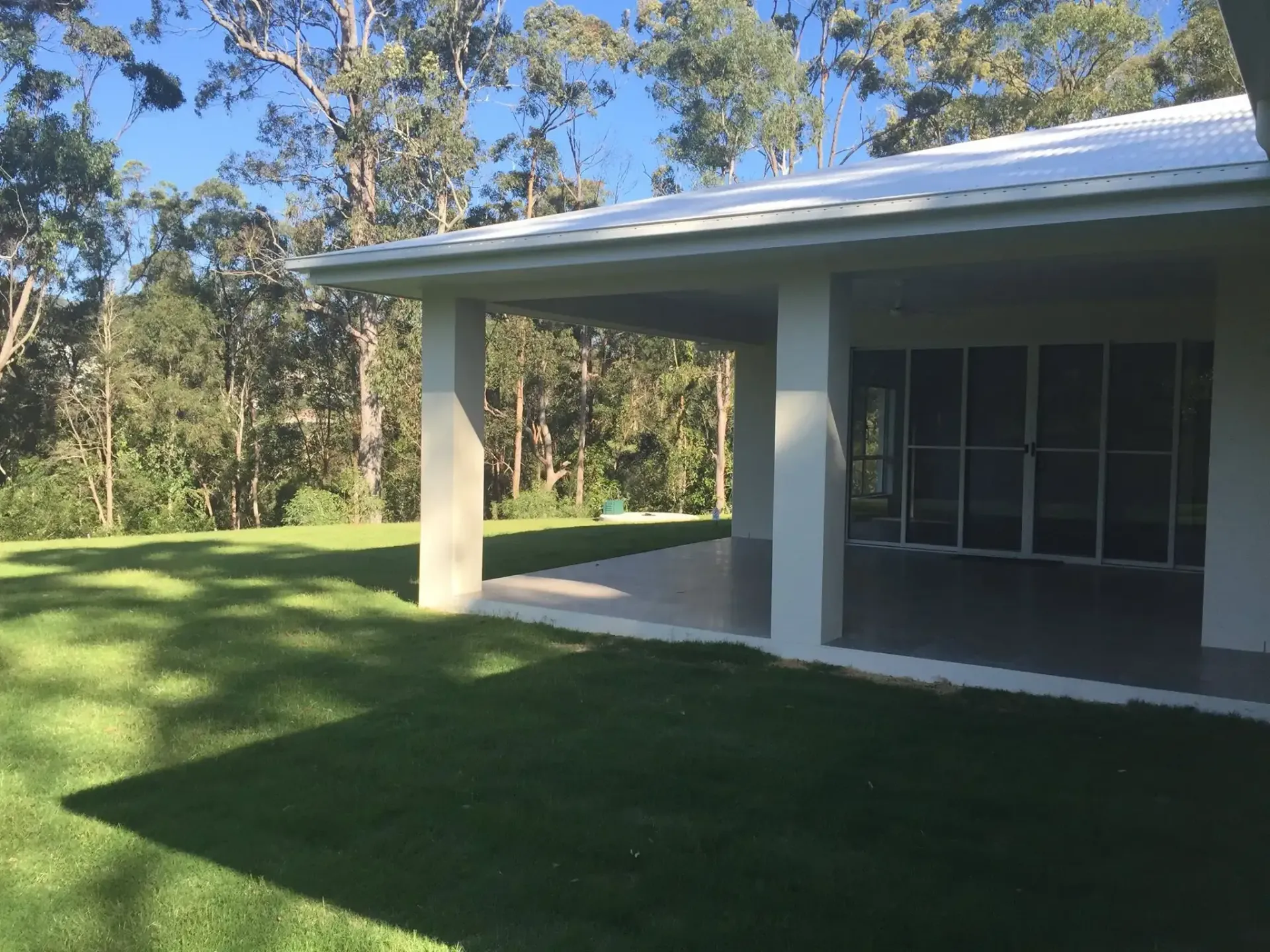 A white, covered patio with large glass sliding doors overlooks a sunny green lawn bordered by a forest of trees. — Michael Duncan Constructions Pty Ltd in Robina, QLD