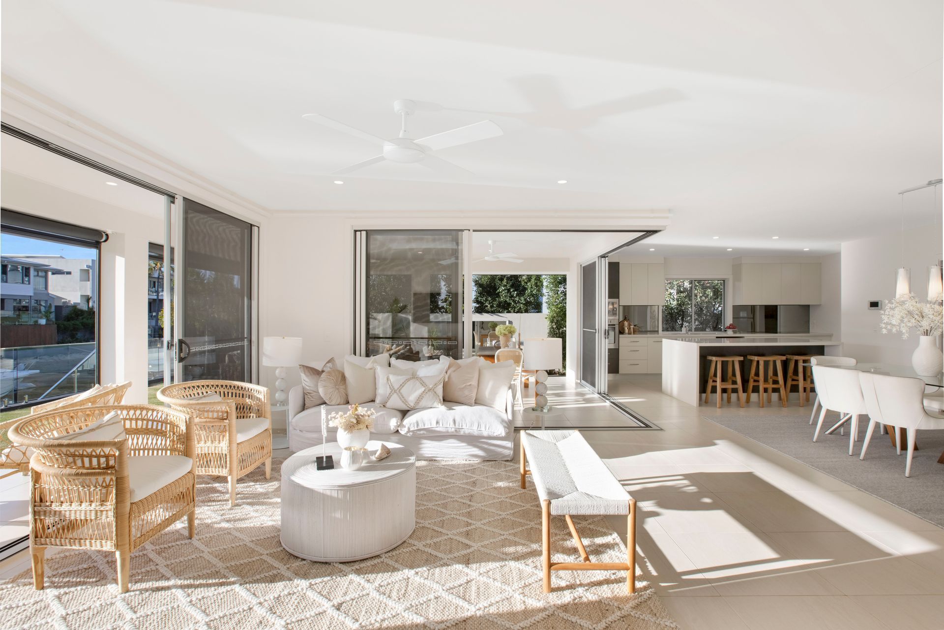A bright, modern open-plan living room with a beige sofa, wicker chairs, a round ottoman, and an adjacent kitchen area — Michael Duncan Constructions Pty Ltd in Mermaid Beach, QLD