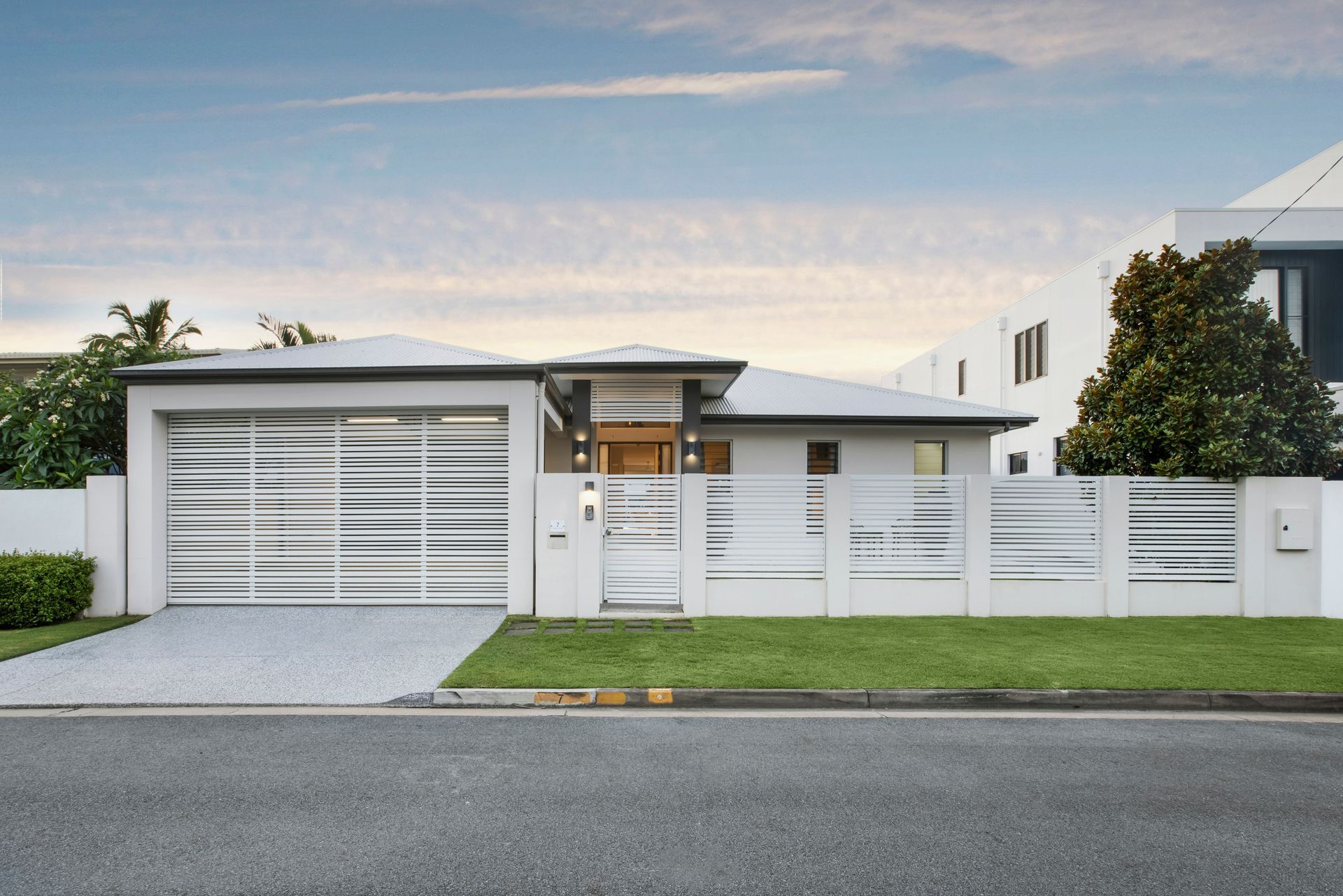 A modern, single-story house with a white horizontal-slat gate and matching privacy fence, front lawn, and gravel driveway — Michael Duncan Constructions Pty Ltd in Robina, QLD