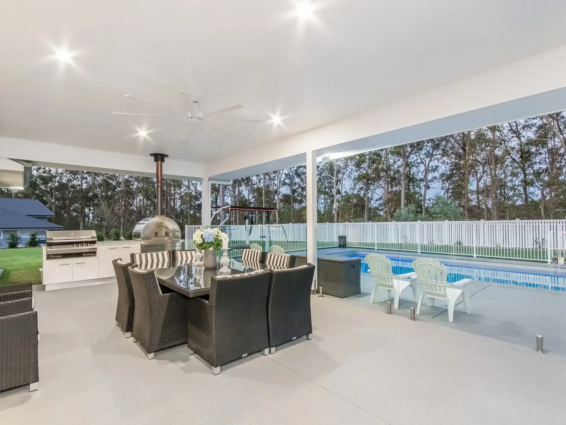 A modern outdoor covered patio with a dark wicker dining set, a pizza oven, and a glass-fenced pool in the background.  — Michael Duncan Constructions Pty Ltd in Robina, QLD