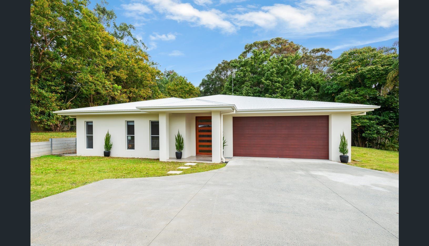 A single-story, white modern house with a wooden front door, double garage, and a large concrete driveway amid greenery — Michael Duncan Constructions Pty Ltd in Robina, QLD