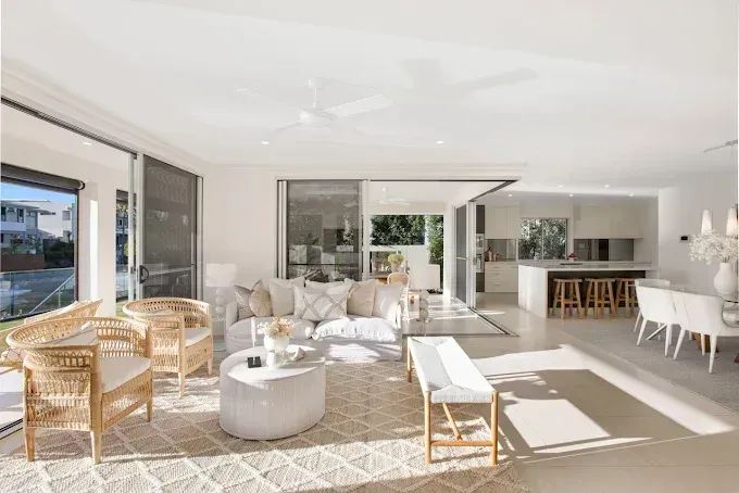 A bright, modern open-plan living room with white decor, woven armchairs, a central sofa, and an adjoining kitchen. — Michael Duncan Constructions Pty Ltd in Robina, QLD