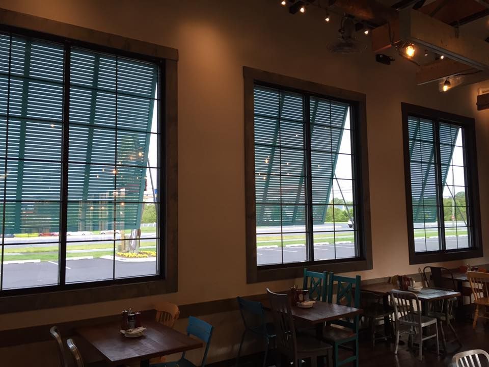 Restaurant interior with three large windows covered with green shades; tables and chairs visible.