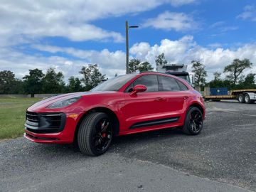 Red Porsche Macan SUV parked on a paved lot with a cloudy sky in the background. Red Porsche Macan SUV parked on a paved lot with a cloudy sky in the background.