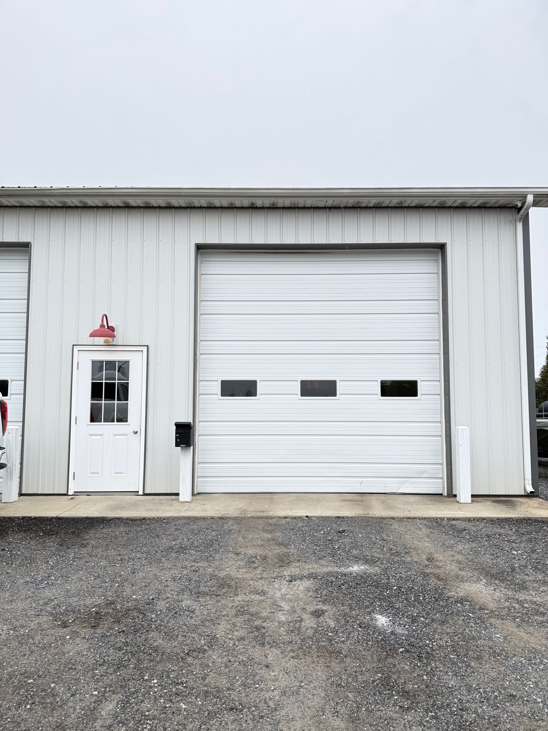 White building with a closed garage door, small door, and gravel ground under an overcast sky.