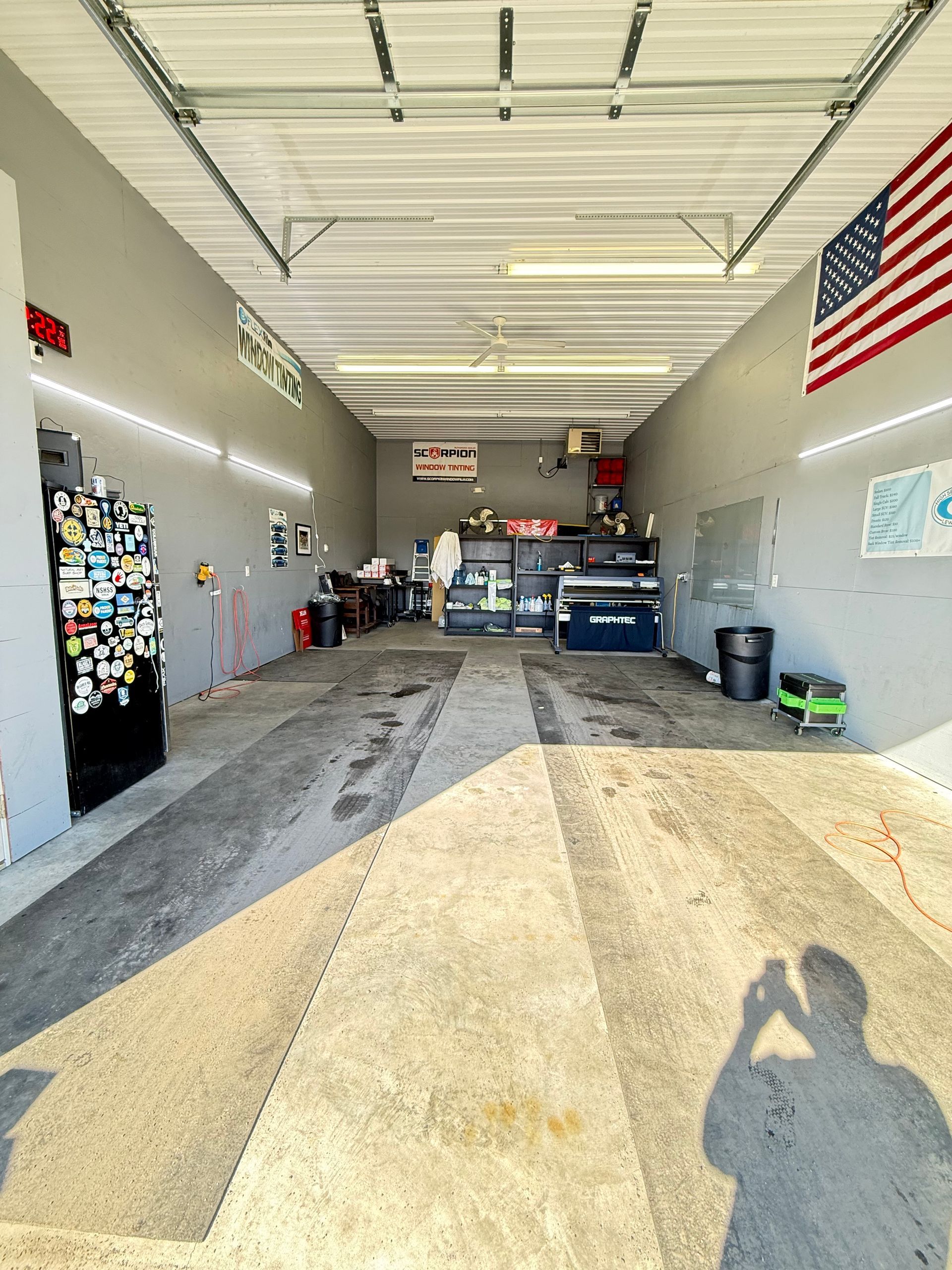 Inside a garage. Concrete floor, two bays, tools and equipment visible. American flag on the wall.