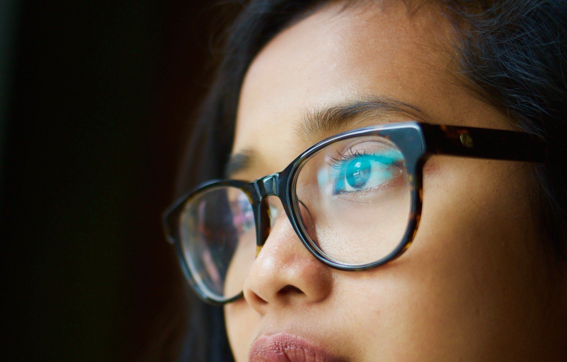 Woman wearing glasses, looking upwards. Black frames, brown skin, dark background.