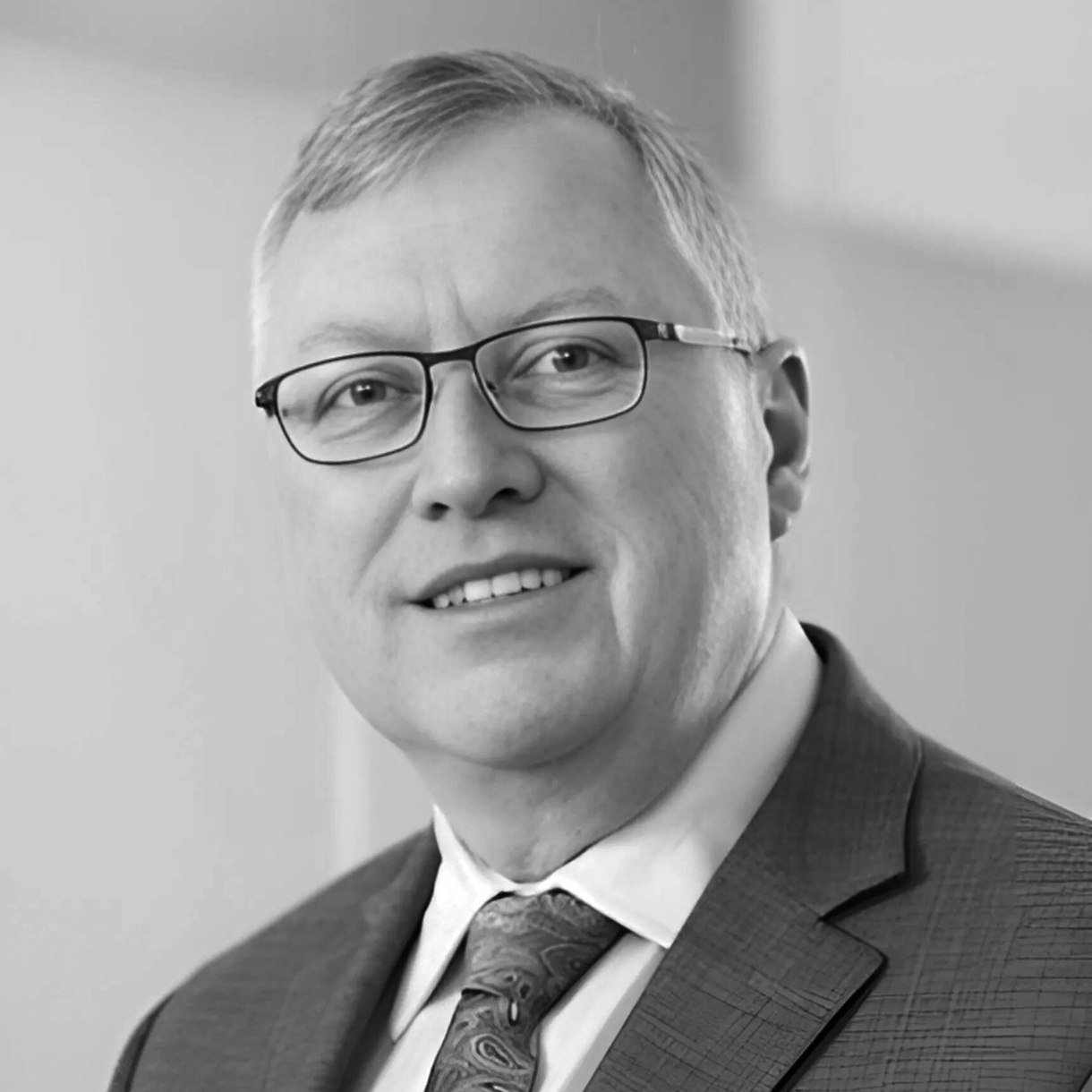 Man in glasses, suit, and tie, looking at the camera, headshot in grayscale.