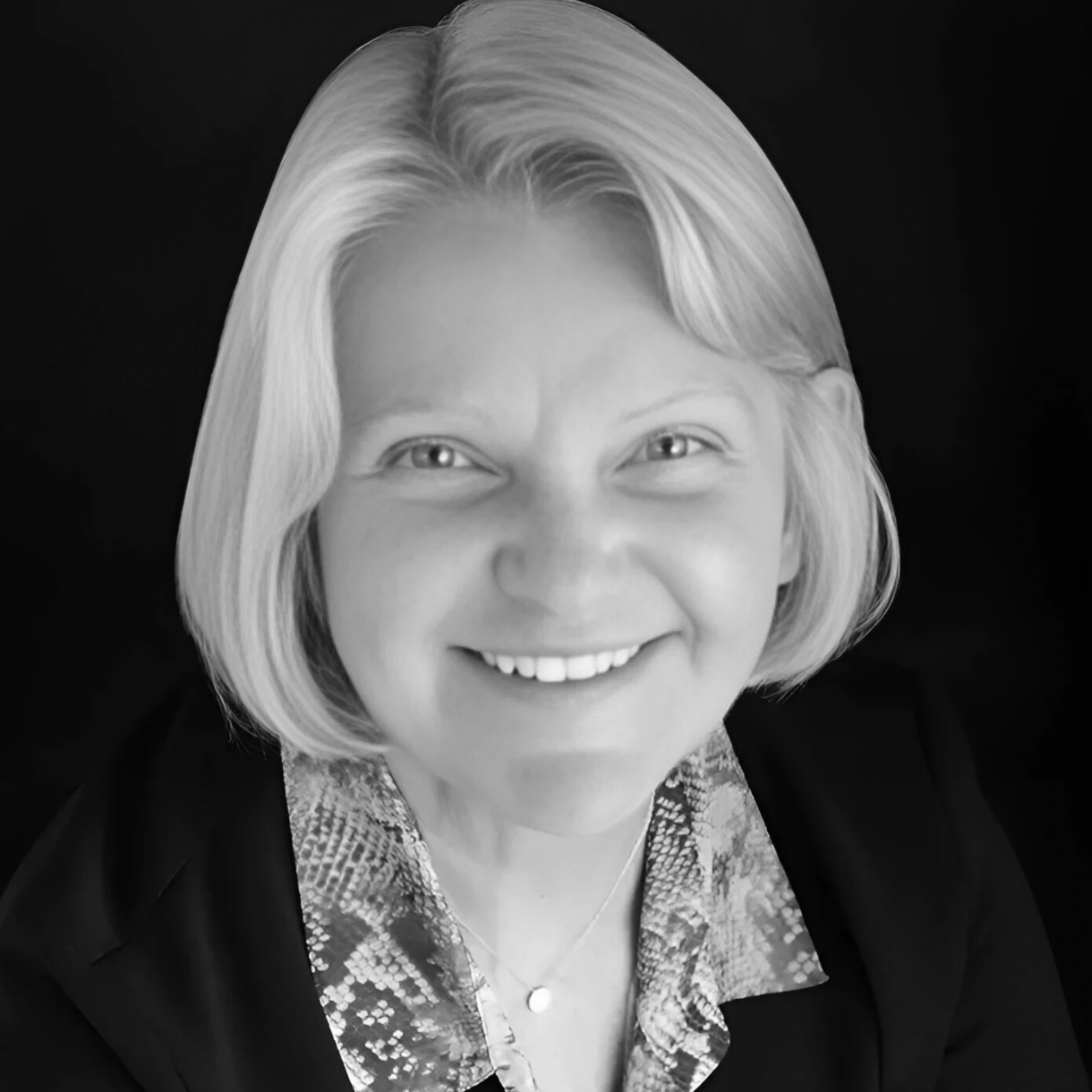 Woman with short, light hair smiles, wearing a blazer over a patterned top, in a studio setting.