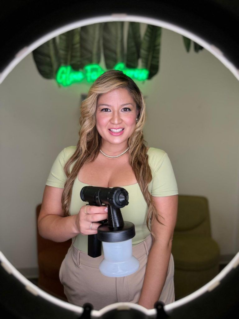 Woman holding a spray gun smiles at the camera, wearing light green top, tan pants, in a studio with a neon sign.