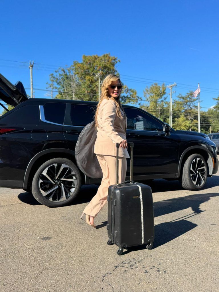 Woman in tan suit rolling a suitcase toward a black SUV with open trunk on a sunny day.