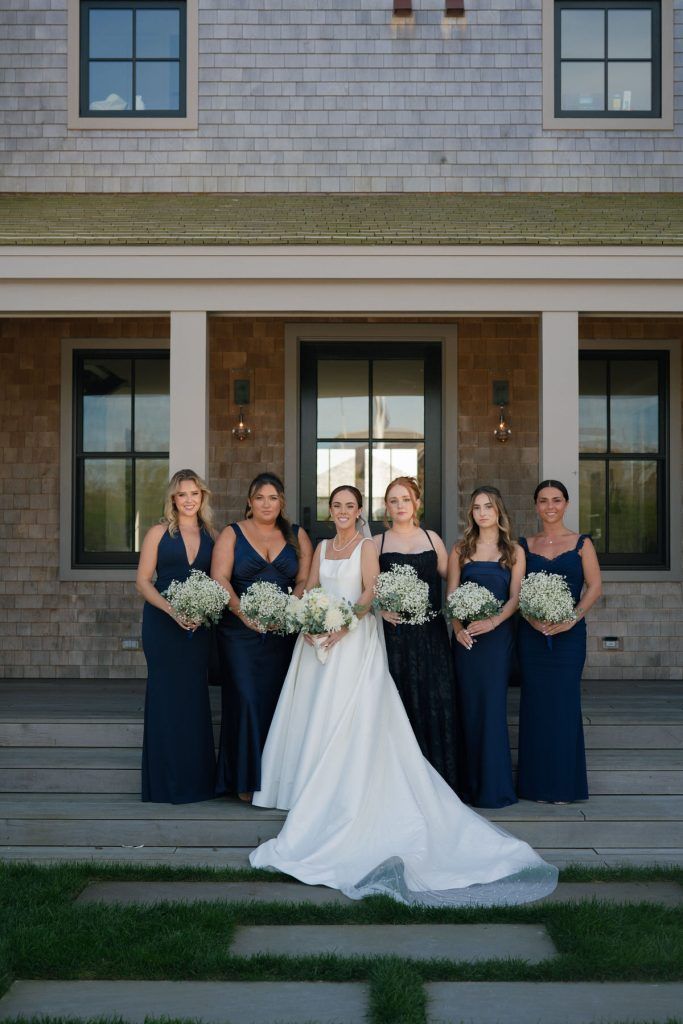 Bride and bridesmaids pose in front of a house. All wear elegant gowns, hold bouquets.