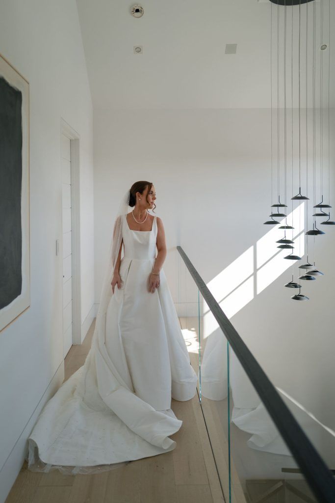 Bride in wedding gown smiles, standing on a bright hallway with modern chandelier.