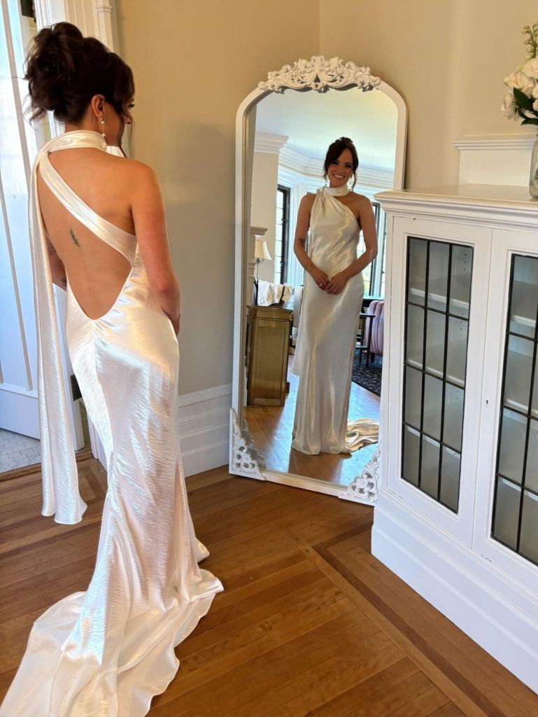 Woman in a white gown with back detail poses in front of a mirror. Interior setting, hardwood floor.