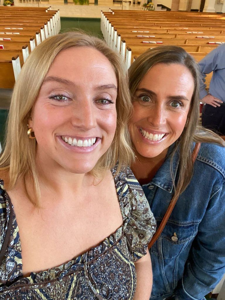 Two smiling women in a church, one in a patterned top, the other in a denim jacket. Rows of pews behind them.