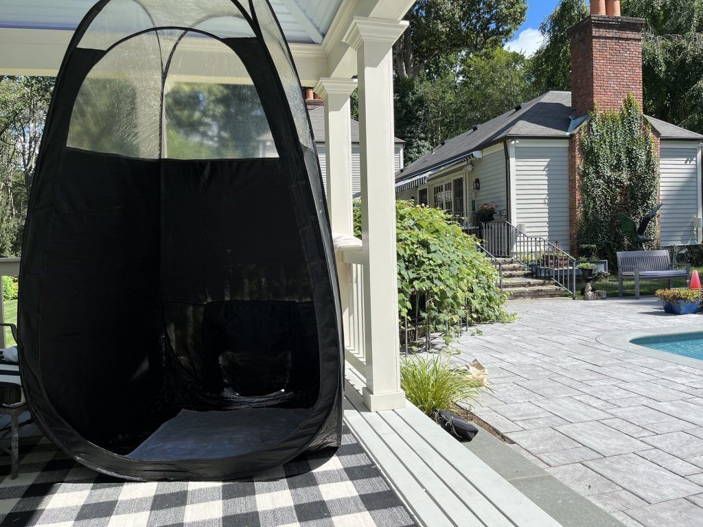 Black spray tan pop-up tent on a checkered rug, porch setting with a house and pool in the background.