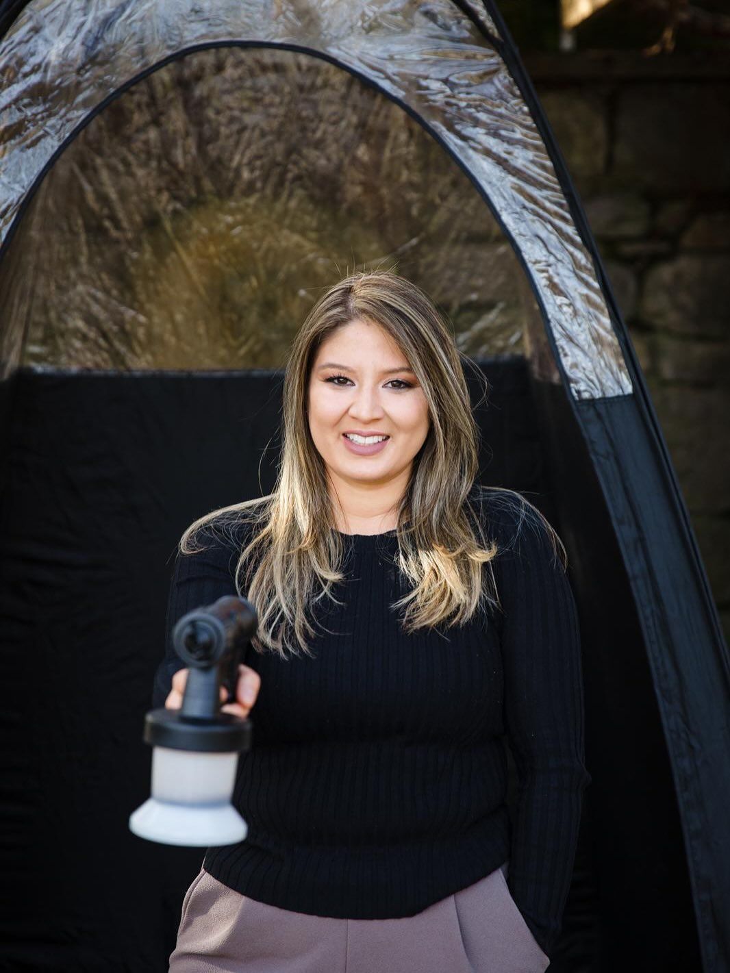 Woman holding spray tan gun, standing in front of a spray tan booth.