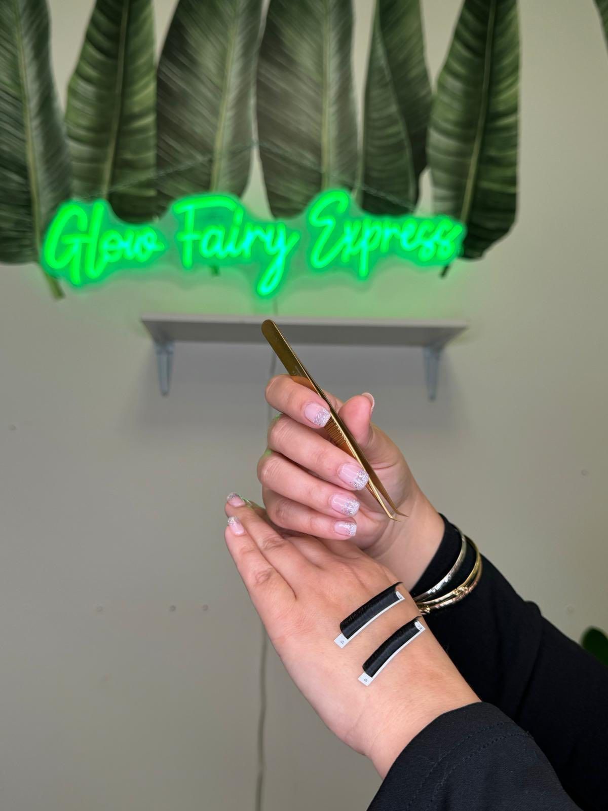 Person holding gold tweezers, next to a black and silver cuff, under a neon sign that reads 