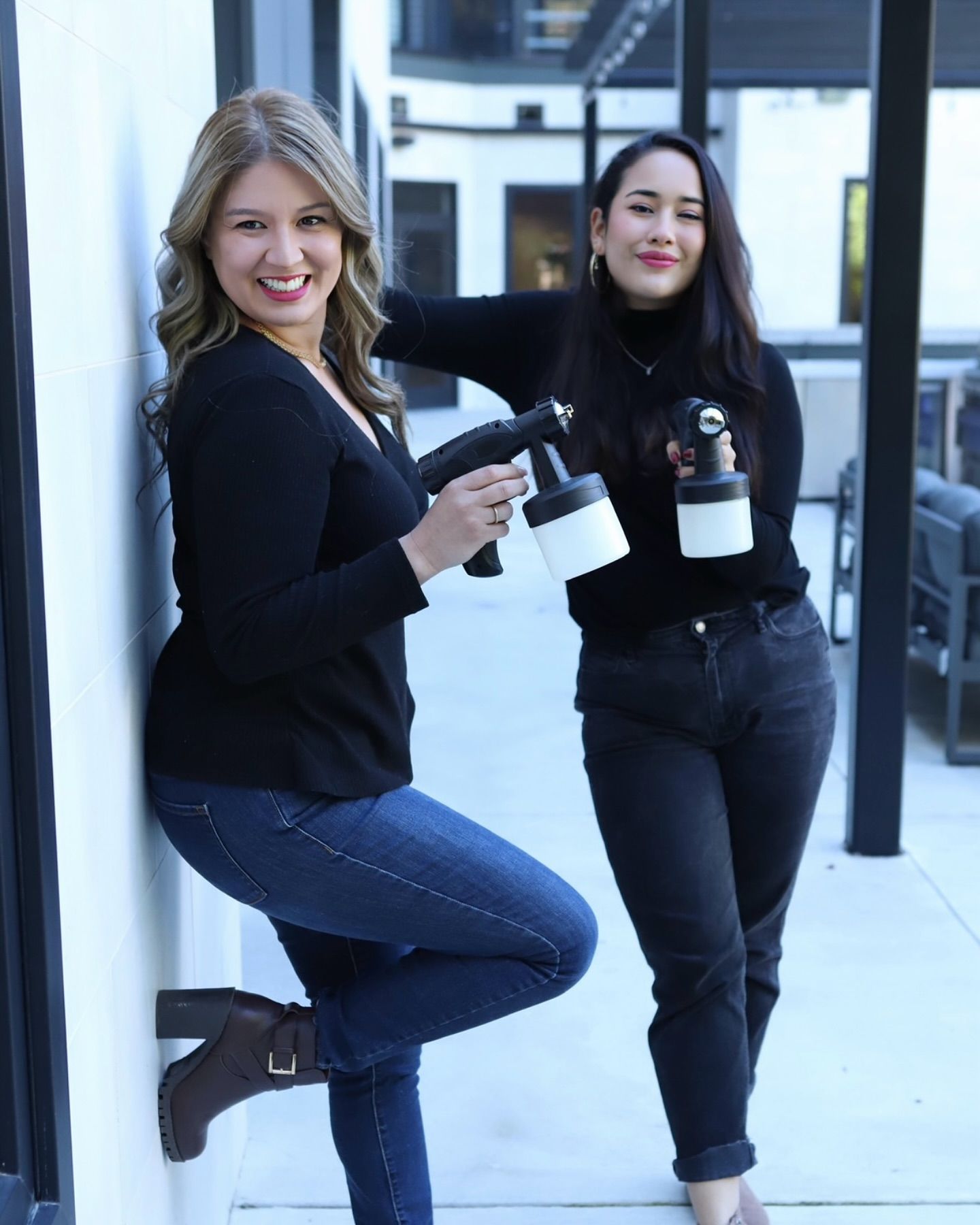 Two women holding spray tan guns smiling in an outdoor setting.