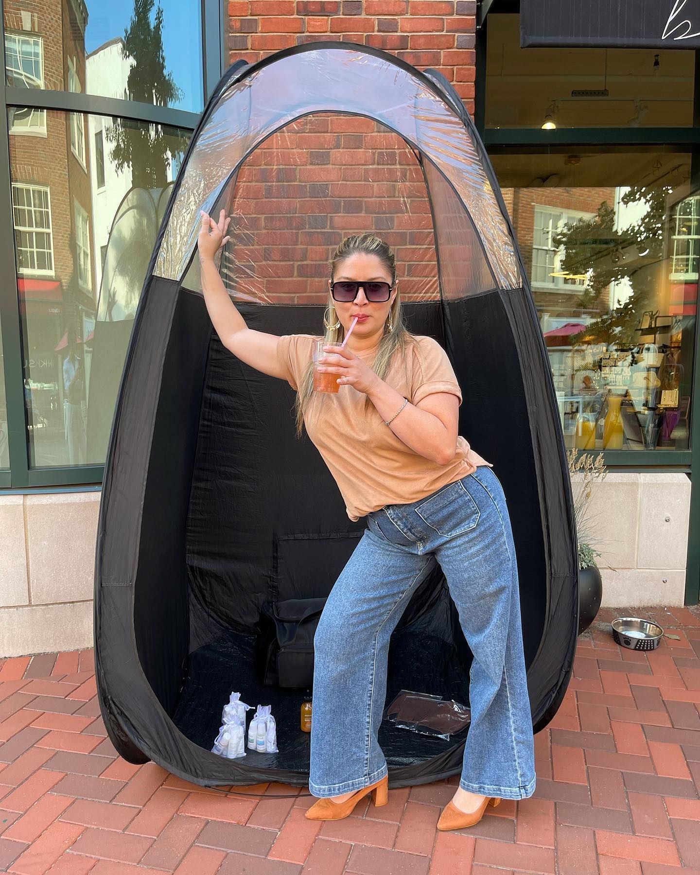 Woman posing in a pop-up spray tan booth outdoors. She wears sunglasses, denim pants, and a tan top.