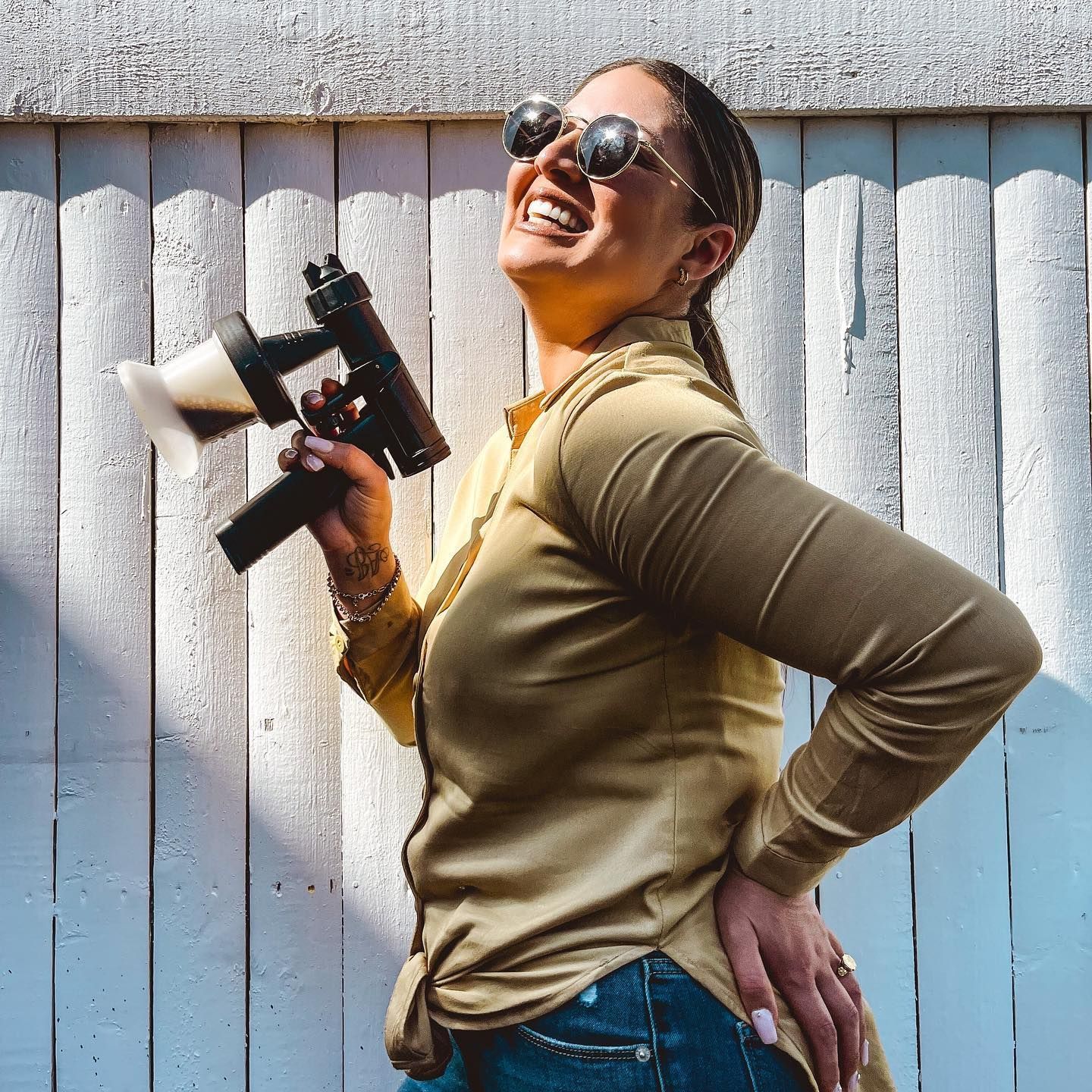 Woman in sunglasses holds a spray tan gun, smiling, leaning against a white fence.