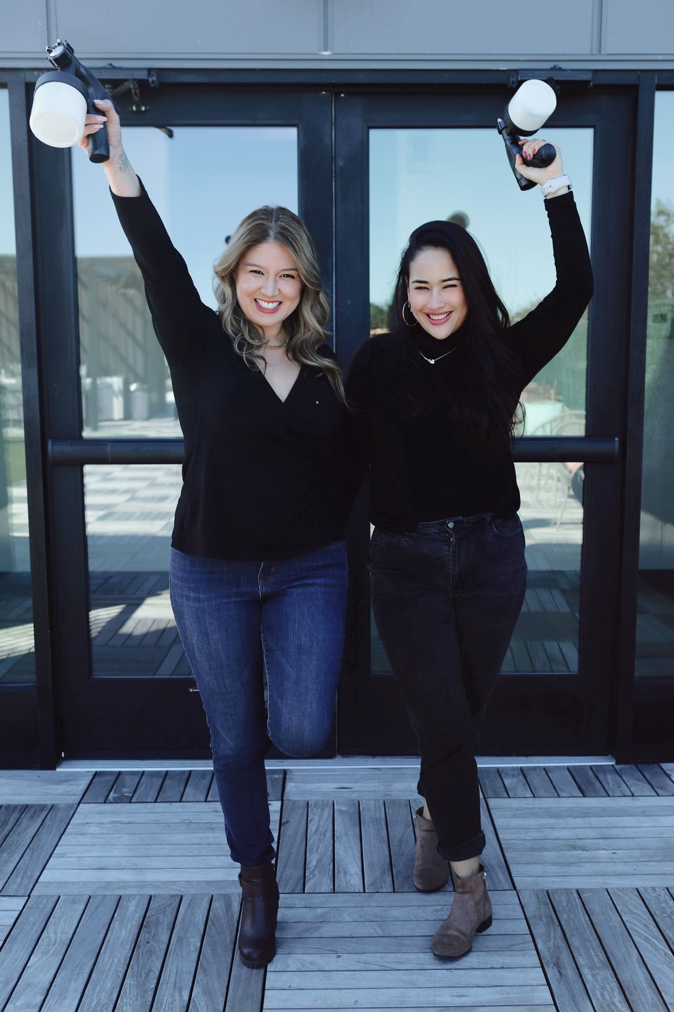 Two women holding spray tan guns with arms raised in front of a glass door.