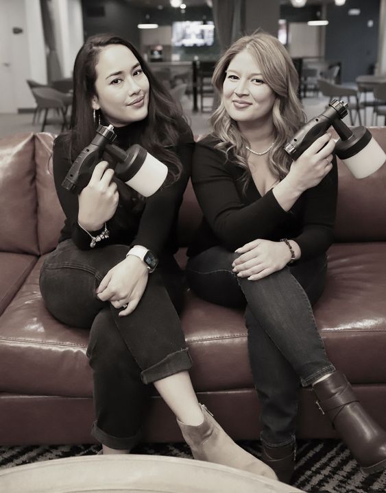 Two women seated on a couch holding bottles, posing indoors and smiling at the camera.