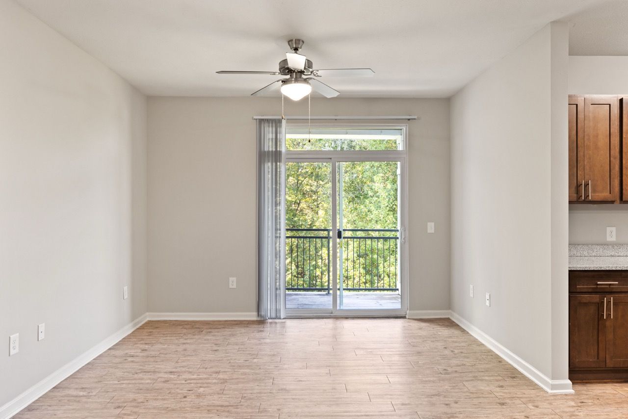Empty living room with sliding glass door to balcony, ceiling fan, and light wood flooring.