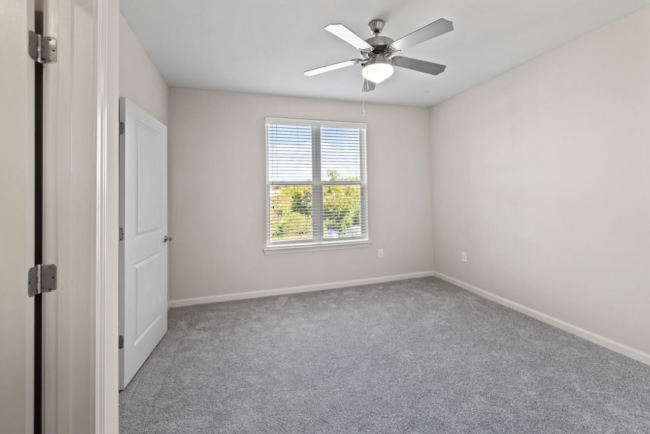 Empty bedroom with gray carpet, light walls, a window with blinds, and a ceiling fan.