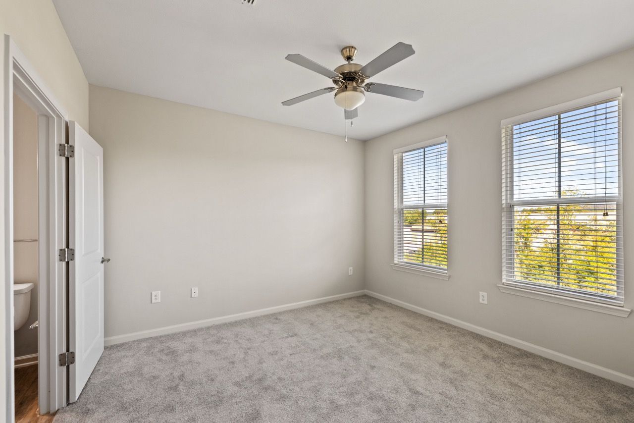 Empty bedroom with beige walls, two large windows with blinds, carpet, and a ceiling fan.