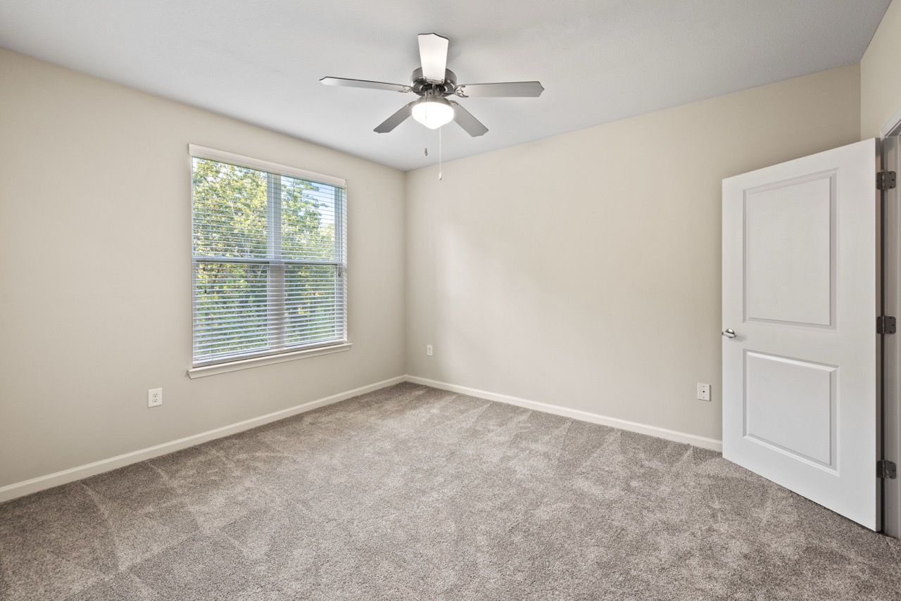 Empty bedroom with window, blinds, ceiling fan, and beige walls.