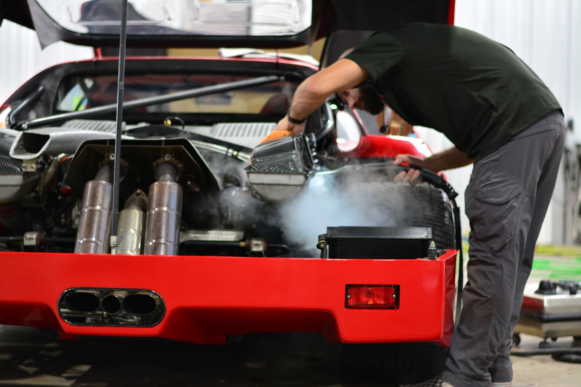 Man steam-cleans a red car engine bay, steam rising.