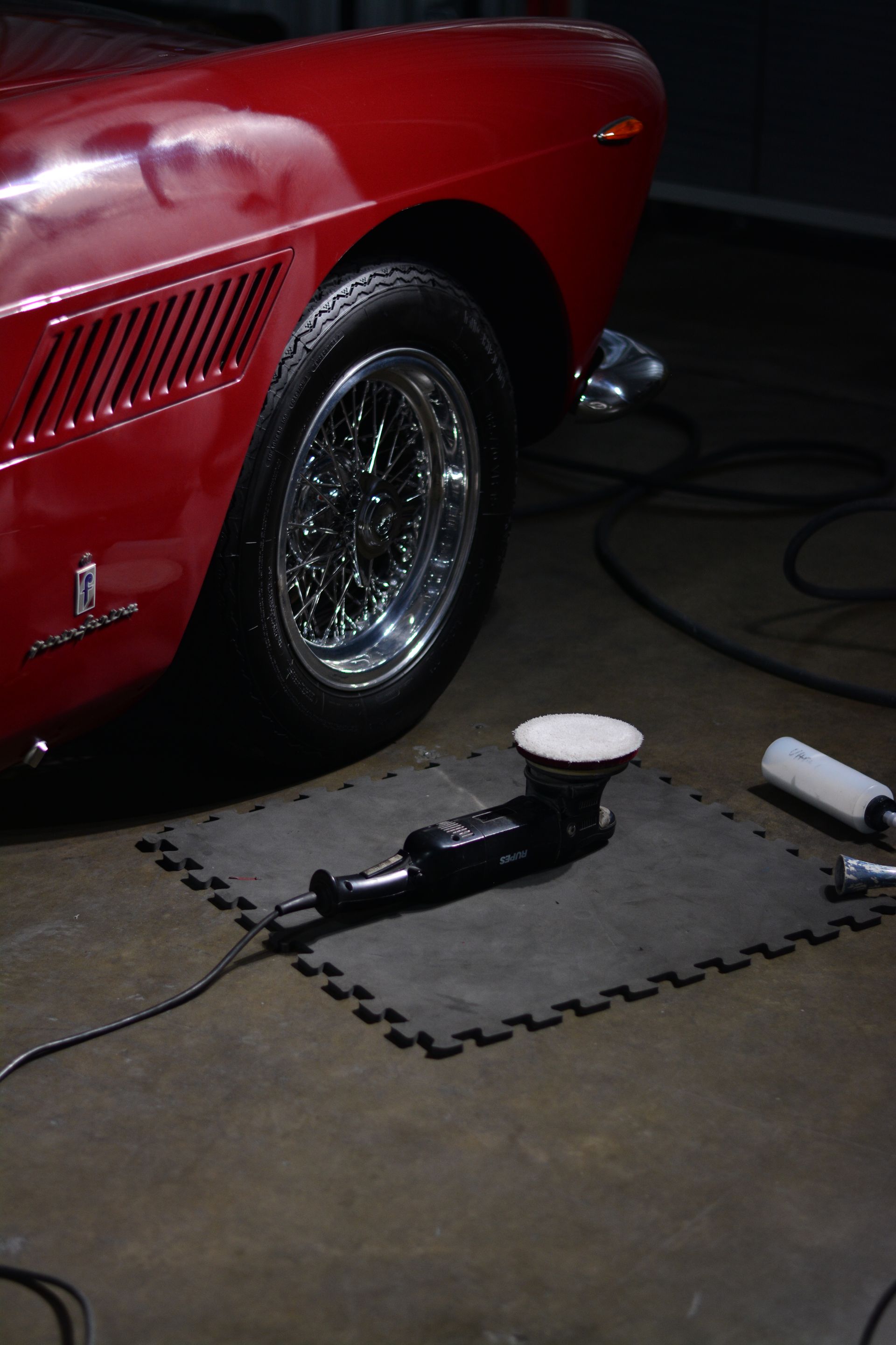 Red classic car being polished, with a detailing machine on a mat. Shiny chrome wheels.