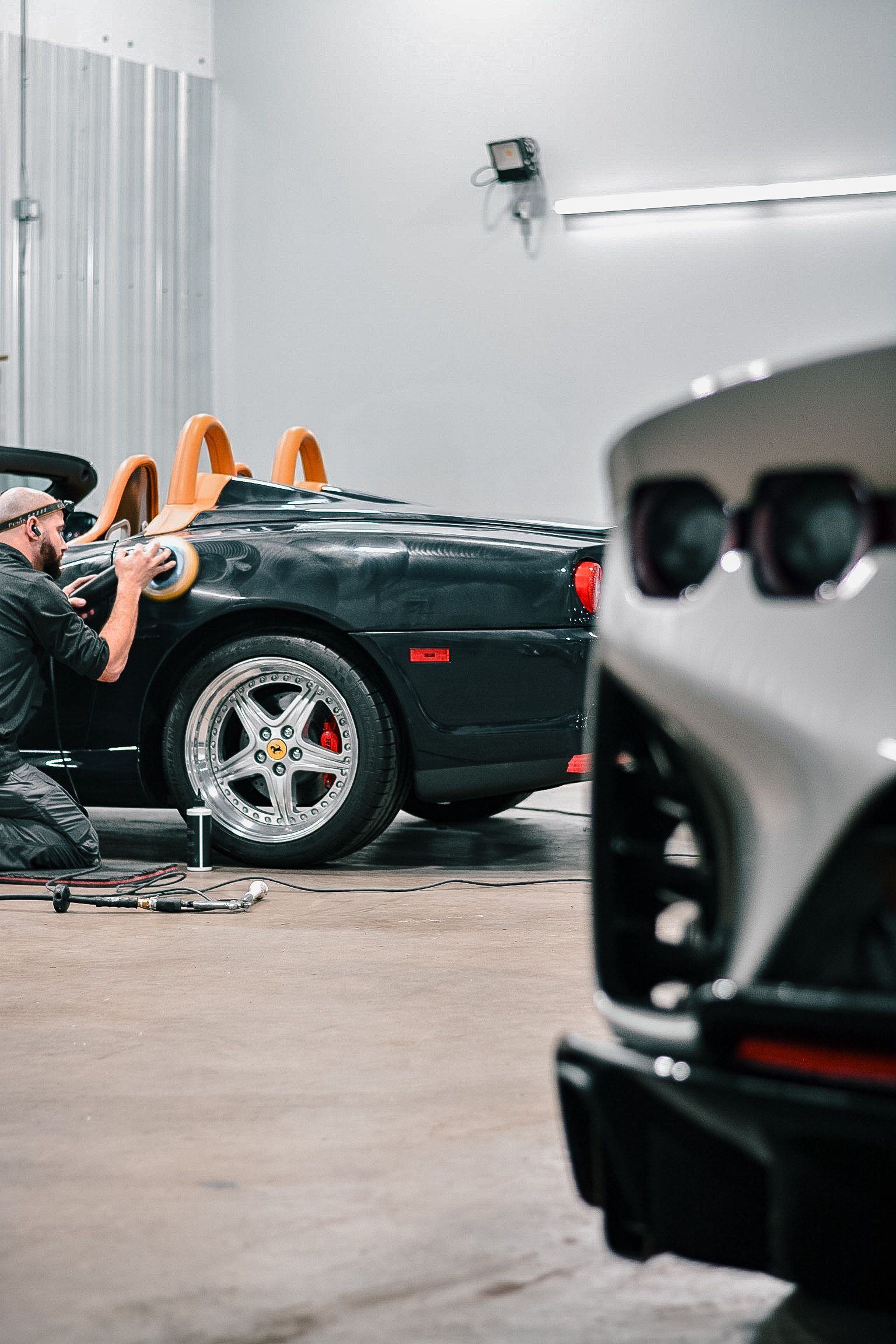 Man polishing a black convertible sports car in a garage with a partially visible silver car in the foreground.