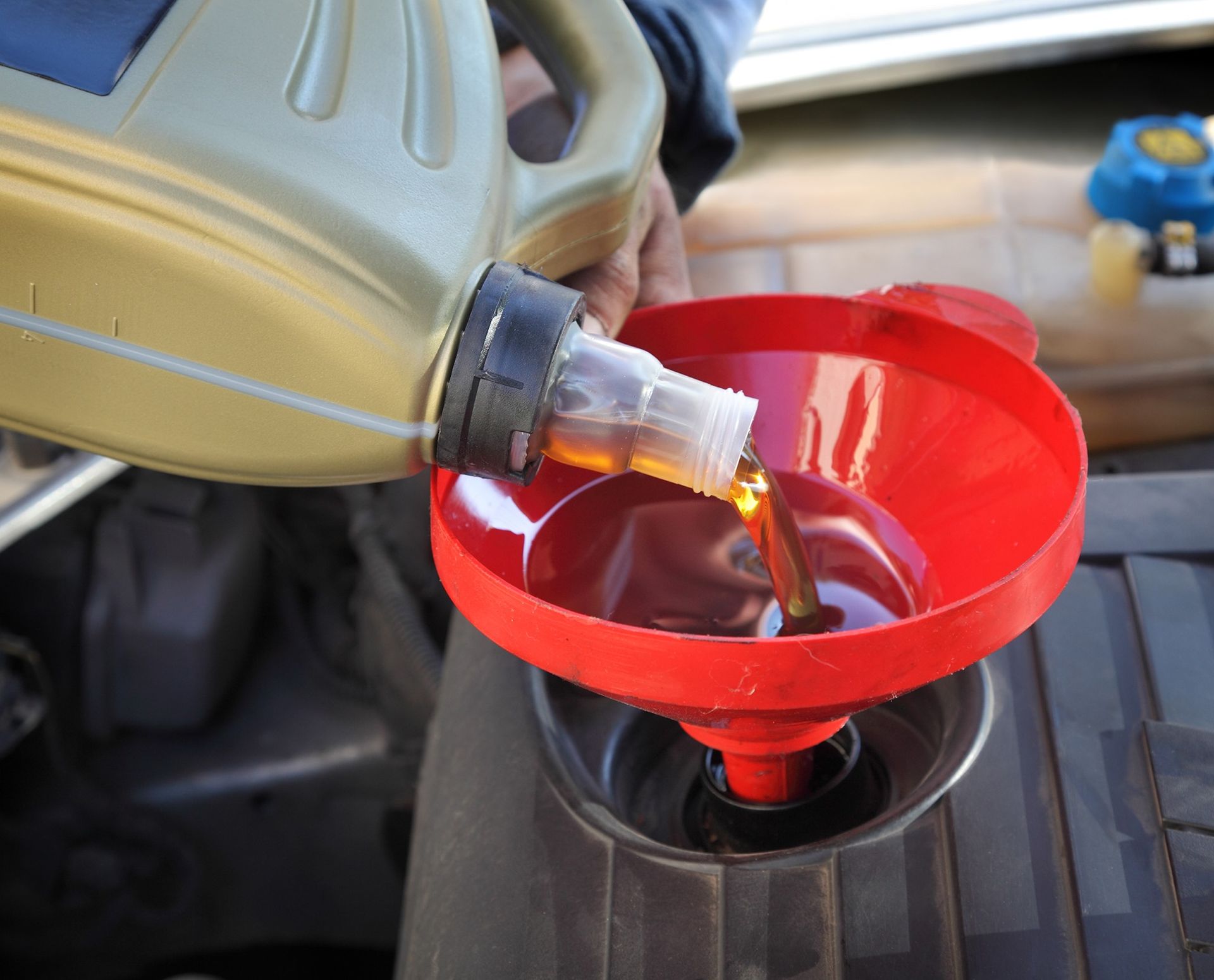 Person pouring oil from a yellow container into a car engine using a red funnel.