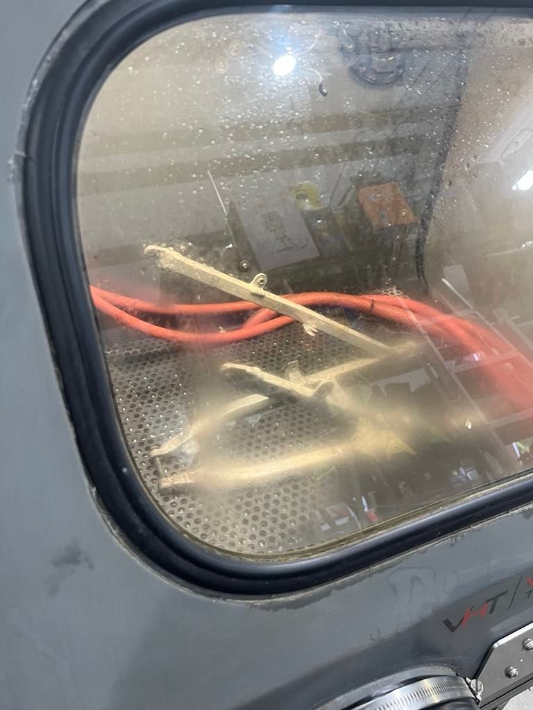 A metal frame and red cables inside a sandblasting cabinet with a glass view window.