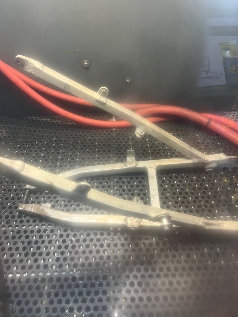 A metal, silver-toned motorcycle swingarm sits on a dark, perforated workshop surface with red cables in the background.