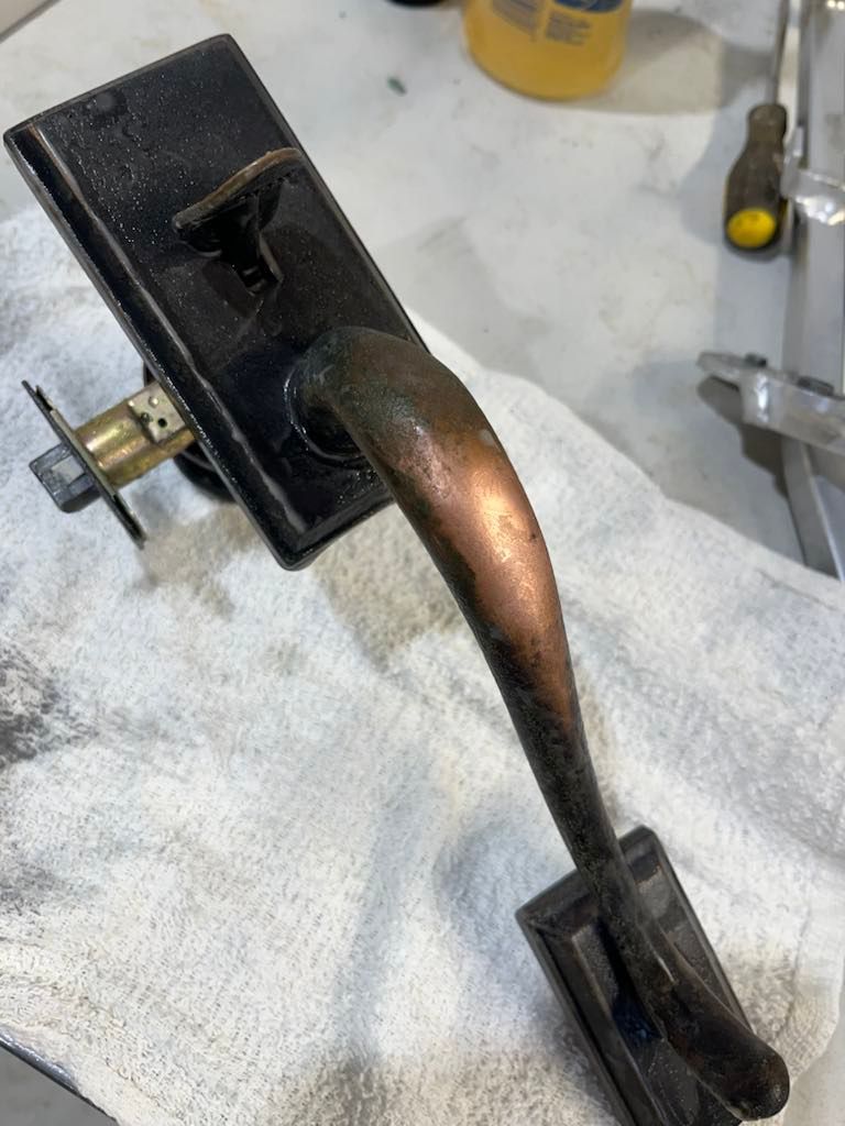 A rustic black door handle with a curved, tarnished copper-toned grip lying on a white cloth surface.