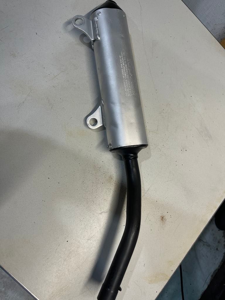 A motorcycle exhaust muffler with a silver cylindrical body and a curved black pipe, viewed from above on a flat surface.