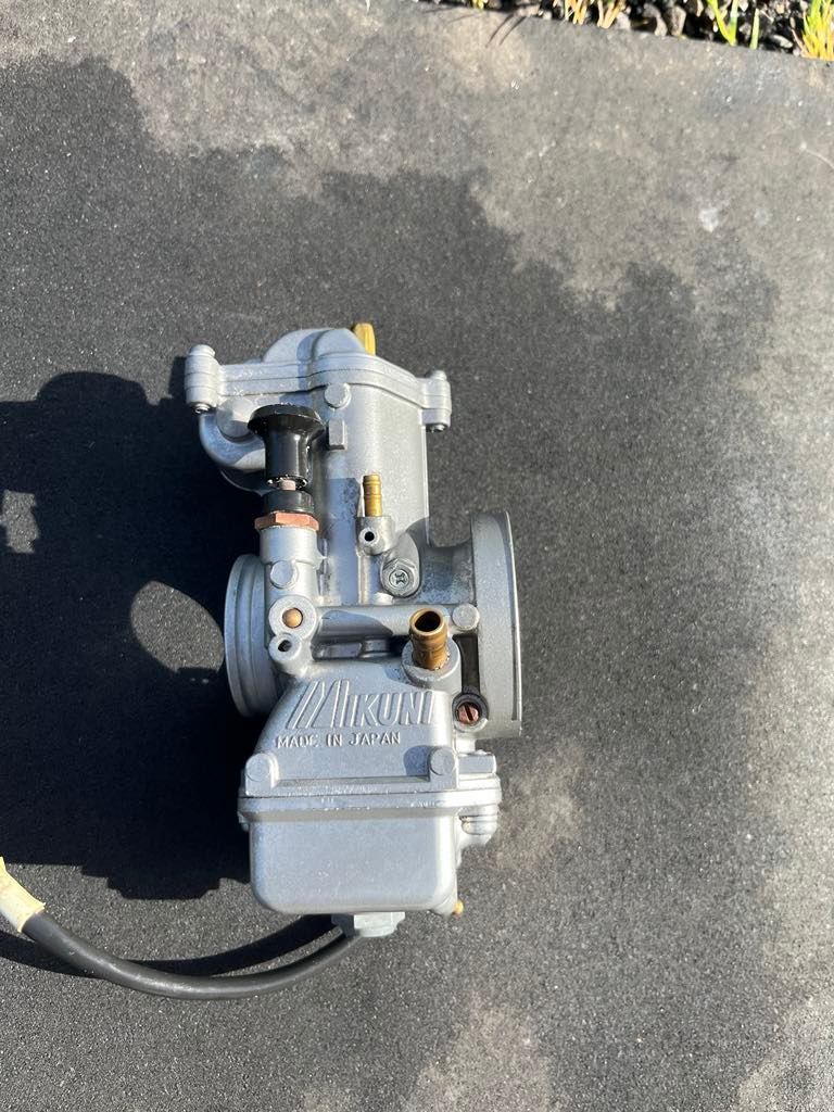 A silver Mikuni carburetor sitting on a dark grey surface, viewed from a top-down angle.