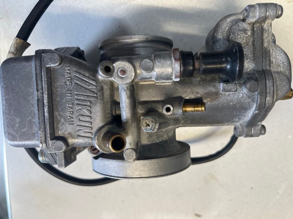 A Mikuni carburetor with a black choke knob and metallic housing, viewed from the side on a light surface.