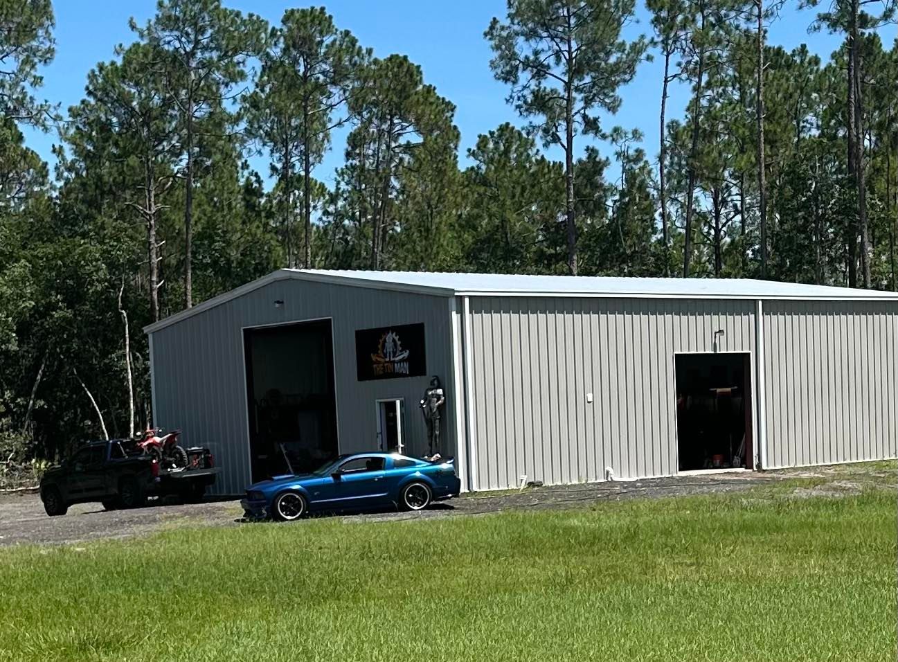 A blue sports car parked in front of a large metal warehouse building surrounded by a dense forest on a sunny day.
