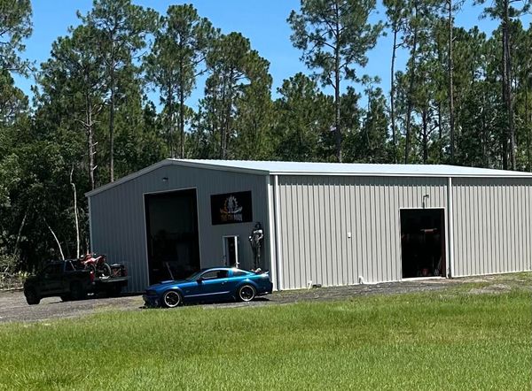 A blue sports car parked in front of a large metal warehouse building surrounded by a dense forest on a sunny day.