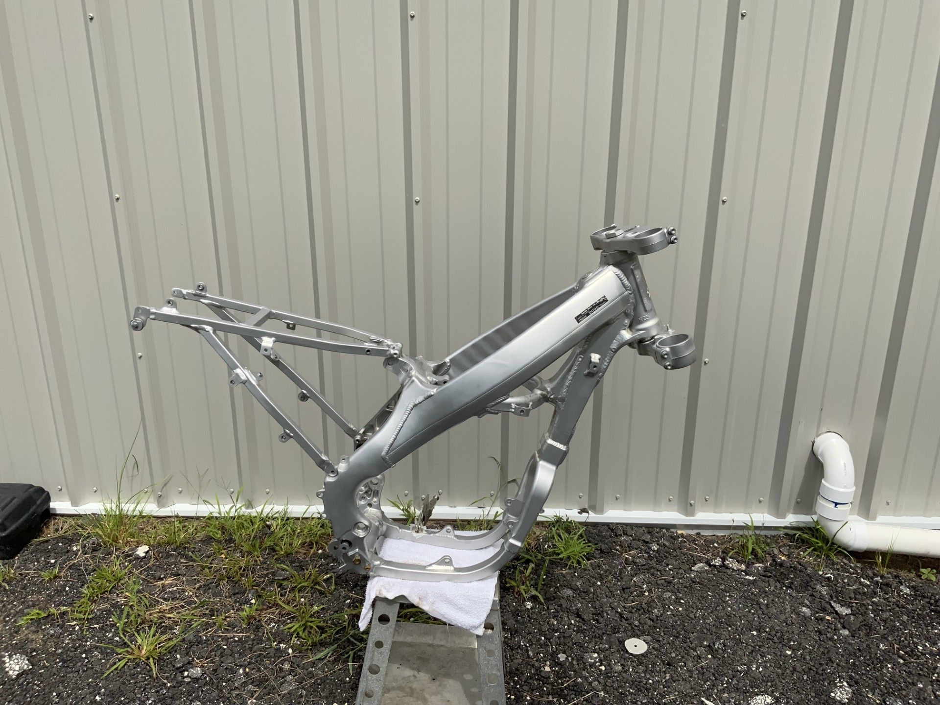 An aluminum motorcycle frame stands on a metal support base in front of a white corrugated metal wall.