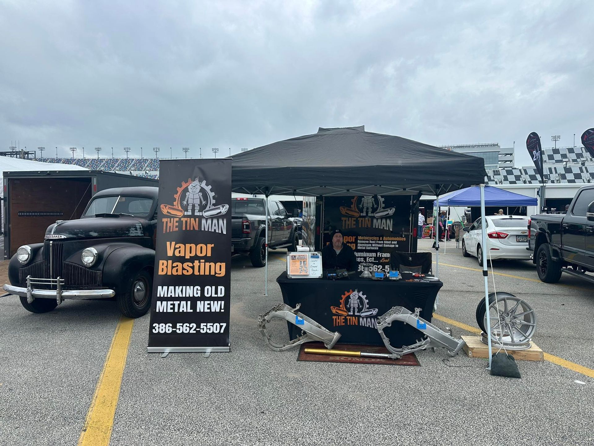 A The Tin Man Vapor Blasting booth with a vintage black car and metal parts on display at an outdoor event.